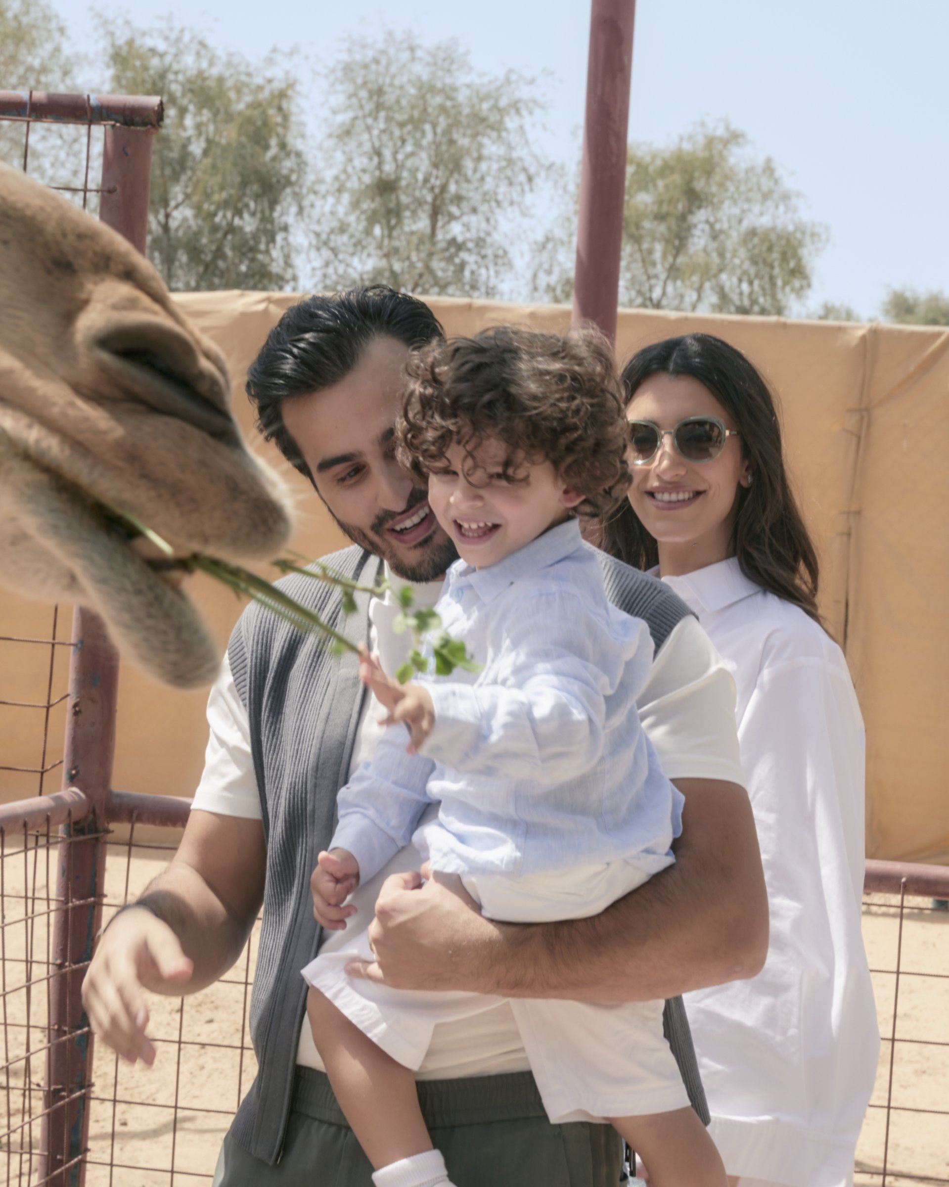 Ali holding Aboudi while feeding a camel, Walaa in the
      background — Emporio Armani campaign by Ali & Walaa, April 2024