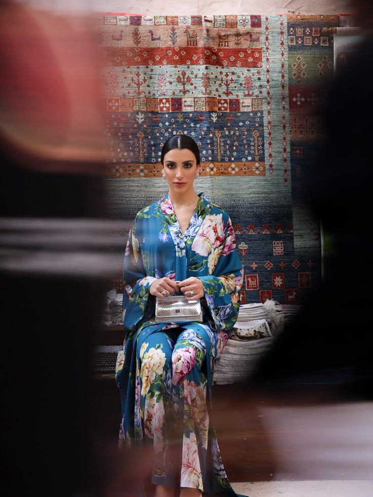 Walaa Awada seated in Dolce & Gabbana Ramadan 2026 teal
      floral silk kaftan at Al Seef, Dubai, framed through a rug
      display — campaign content by Ali & Walaa (@aliandwalaa)