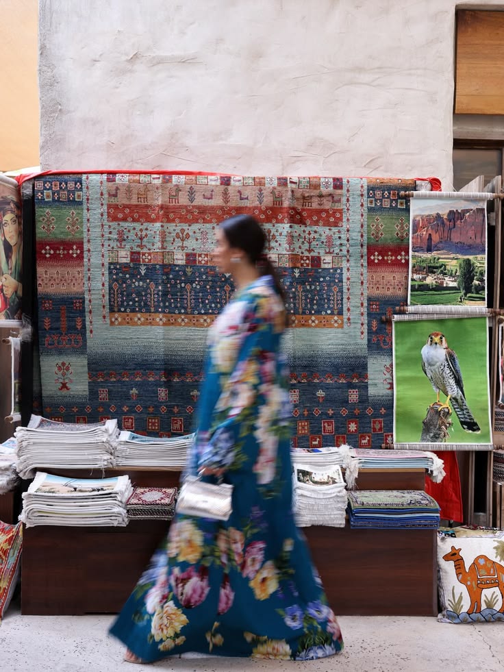 Walaa Awada in motion wearing Dolce & Gabbana Ramadan 2026
    teal floral silk kaftan, walking past the rug displays at
    Al Seef, Dubai — Dolce & Gabbana campaign by Ali & Walaa,
    March 2026