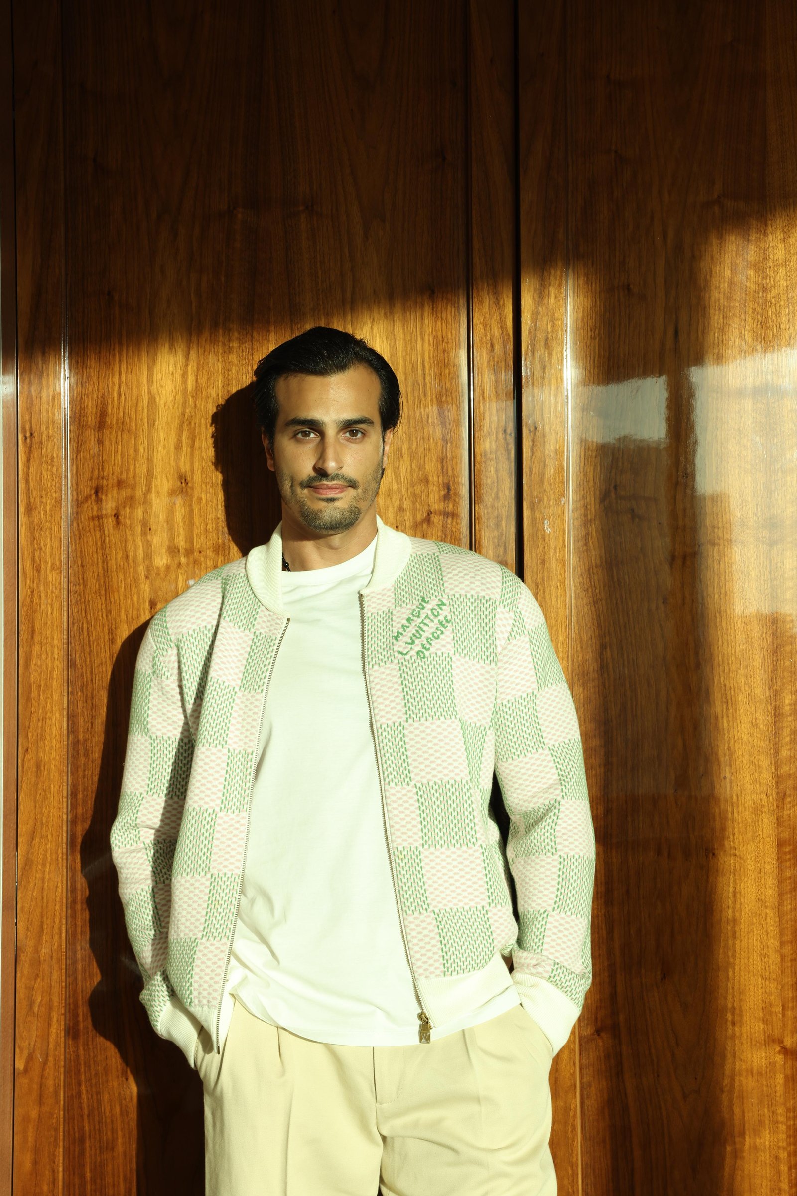 Ali Hennaoui in a Louis Vuitton checkerboard bomber jacket
      against a wood-paneled door at The Lana, Business Bay, Dubai —
      Louis Vuitton campaign by Ali & Walaa, May 2025