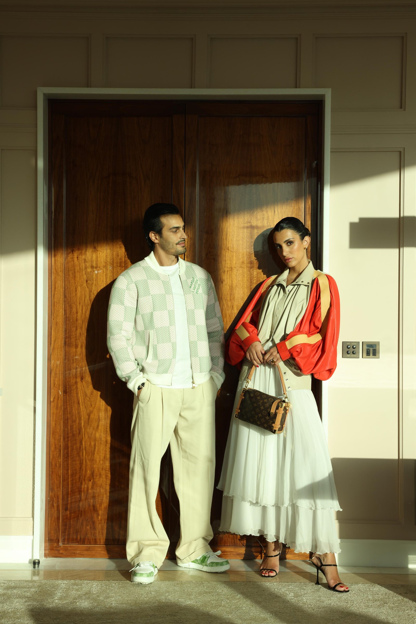Ali Hennaoui and Walaa Awada in Louis Vuitton checkerboard
    jacket and colorblock leather jacket with pleated skirt, standing
    before a wooden door at The Lana hotel, Business Bay, Dubai —
    Louis Vuitton campaign by Ali & Walaa, May 2025