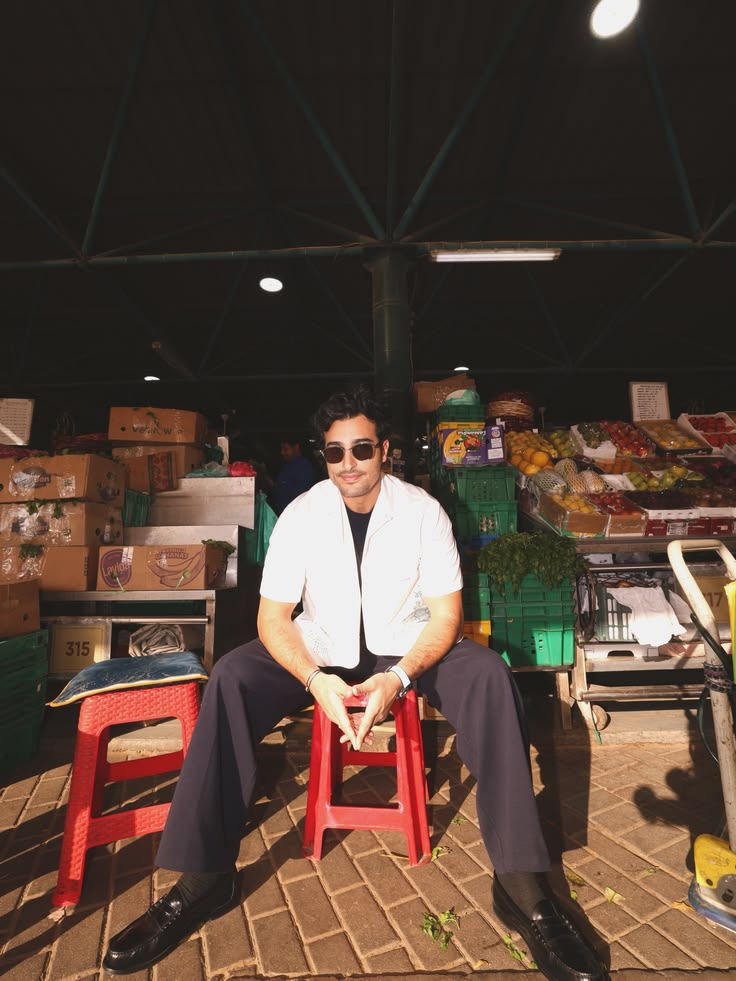 Ali Hennaoui in Prada white graphic-print shirt sitting
      on a red stool at Ras Al Khor market Dubai — Prada SS26
      campaign by Ali & Walaa (@aliandwalaa)