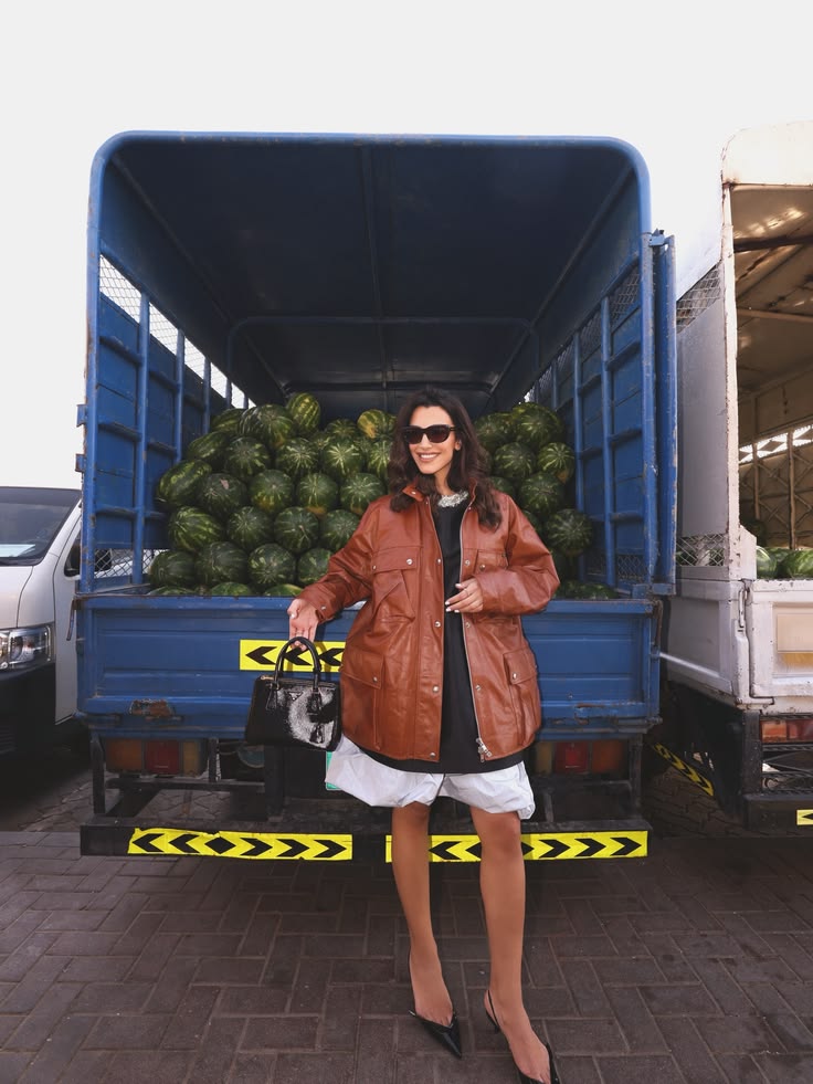 Walaa Awada in Prada Spring-Summer 2026 tan leather jacket
    and Galleria bag standing in front of a watermelon delivery truck
    at Ras Al Khor market Dubai — Prada campaign by Ali & Walaa 2026
