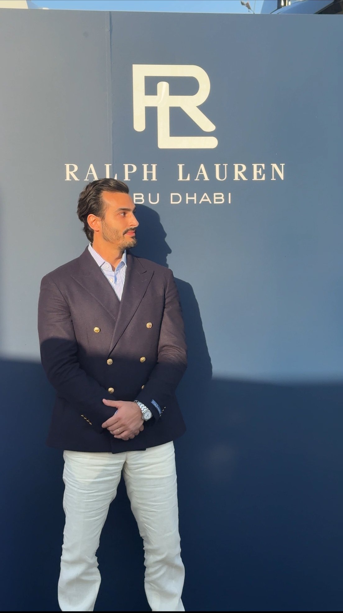 Ali Hennaoui in a double-breasted blazer in front of the
      Ralph Lauren Abu Dhabi sign — Ralph Lauren campaign by Ali &
      Walaa, December 2024