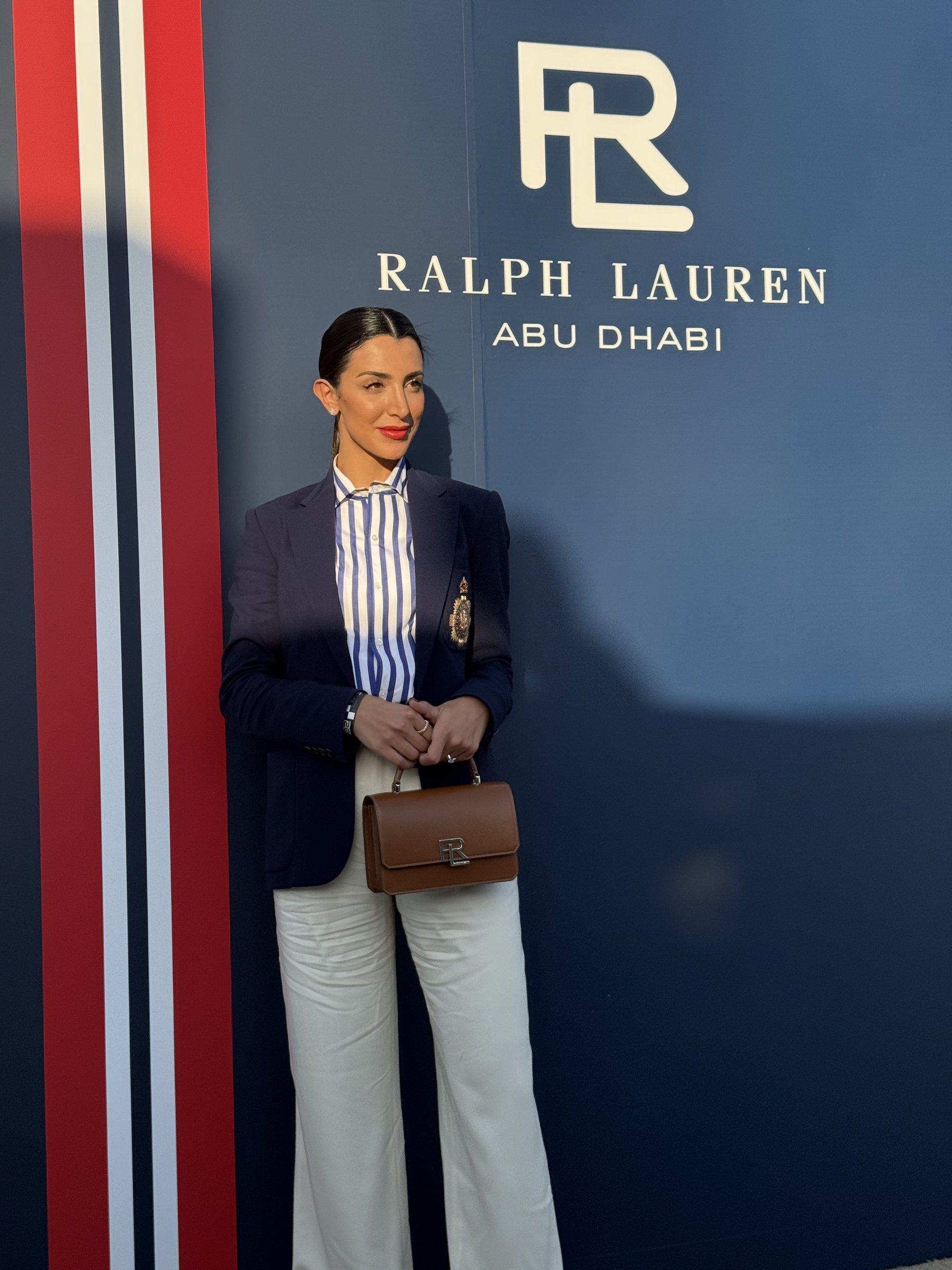 Walaa Awada in a navy crest blazer in front of the Ralph
      Lauren Abu Dhabi sign, holding an RL bag — Ralph Lauren campaign
      by Ali & Walaa, December 2024